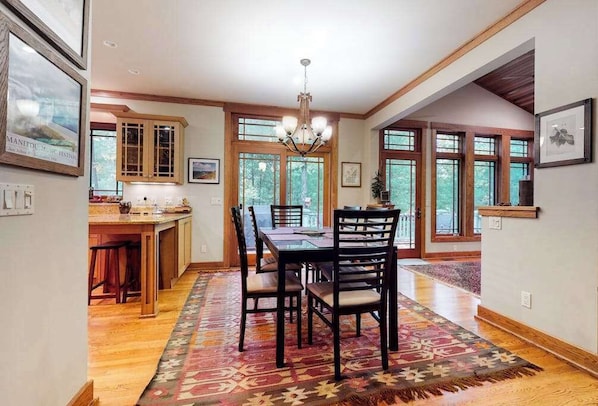 A spacious dining area with a wooden table, chairs, and a chandelier. There are large windows providing natural light and views of the outdoors.
