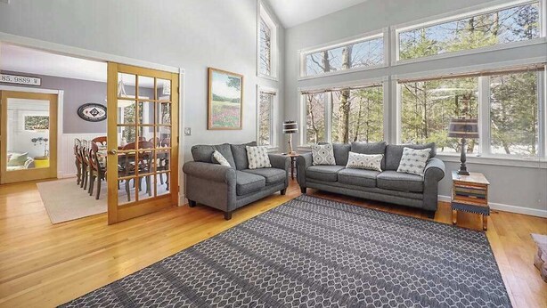 Ein geräumiges Wohnzimmer mit zwei grauen Sofas, einem gemusterten Teppich und großen Fenstern mit Blick auf Bäume.