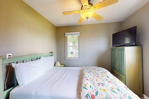 Un dormitorio con una cama, ventilador de techo, televisor sobre un mueble y una ventana.