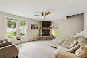 A living room with a fireplace, a ceiling fan, and a large window.