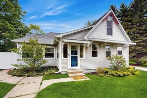 Une maison de plain-pied avec un porche à l’avant, une porte bleue et un petit jardin devant, agrémenté d’un arbre et de quelques arbustes.