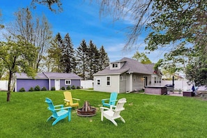 A backyard with a fire pit, three Adirondack chairs, and a house with a sloped roof.