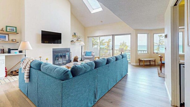 A spacious living area with a large sofa, a flat-screen TV mounted on the wall, a fireplace, and a balcony with large windows. - Darling condo on Lake Michigan with washer/dryer, deck, lake views, fireplace (Glen Arbor)