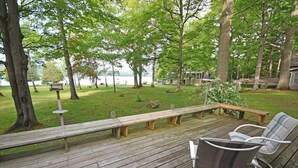 A wooden deck with benches and chairs, surrounded by trees and a lake view. - Lakefront home on Little Glen Lake with fireplace, deck, gas grill, and firepit (Empire)