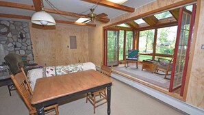 A wooden dining table with chairs, a sofa, and a ceiling fan in a room with large windows and a stone wall. - Lakefront home on Little Glen Lake with fireplace, deck, gas grill, and firepit (Empire)
