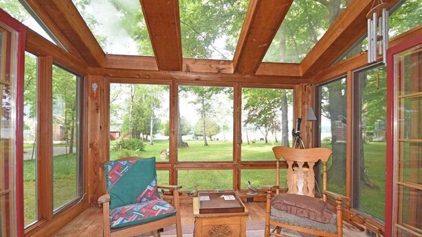 A wooden-paneled room with large windows, a rocking chair, a table, and a view of a green landscape. - Lakefront home on Little Glen Lake with fireplace, deck, gas grill, and firepit (Empire)
