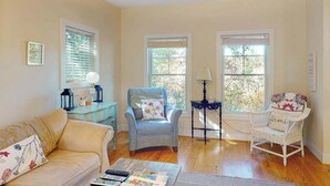Un salón con un sofá, dos sillones, una mesa de centro y una mesita de noche con una lámpara. Tres ventanas dejan entrar luz natural.