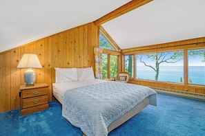 A bedroom with a large bed, a nightstand, a lamp, and a view of the sea through large windows.