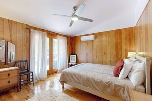 A bedroom with wooden walls, a bed, a desk, a chair, a ceiling fan, and a window with curtains.