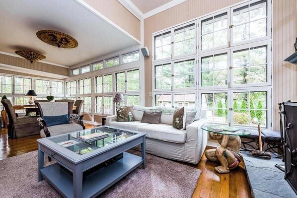 A spacious living room with large windows, a glass coffee table, a white sofa, and a fireplace. - Secluded, charming home with in-ground pool, hot tub, firepit & high-speed WiFi (Union Pier)