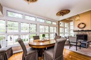 A spacious room with large windows, a round wooden table, and a fireplace. - Secluded, charming home with in-ground pool, hot tub, firepit & high-speed WiFi (Union Pier)