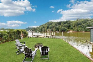 Eine grüne Terrasse mit Adirondack-Stühlen, einem kleinen Tisch und einem Steg, der in einen See führt, umgeben von Bäumen.