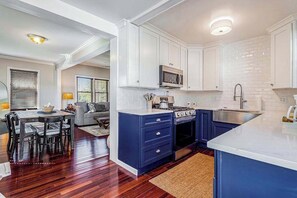 A modern kitchen with white cabinets, a blue island, and a dining area with a wooden table and chairs. - Quaint, budget-friendly home walking distance to town with grill - dog-friendly (Three Oaks)
