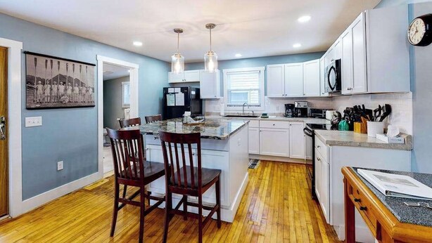 Una cocina con suelo de madera, armarios blancos, zona de comedor con dos sillas, luces colgantes y un reloj en la pared.