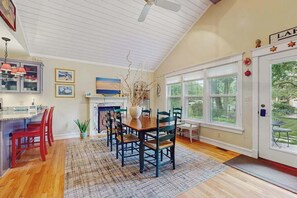 A dining area with a wooden table and chairs, a kitchen with red chairs, and a large window with a view of greenery. - Adorable Downtown Cottage with Firepit, Fireplace, & Yard - Walk Everywhere (New Buffalo)