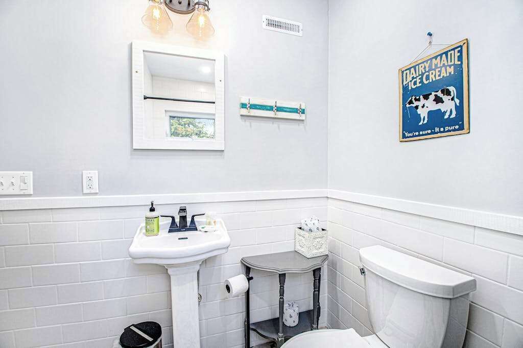 Ein Badezimmer mit einem weißen Waschbecken, Spiegel und Toilette. An der Wand hängt ein gerahmtes Schild und eine kleine Ablage mit Toilettenartikeln.