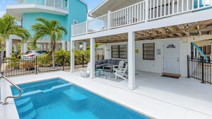 A pool area with a white deck, a covered seating area with a sofa and chairs, and a two-story building with a balcony in the background.