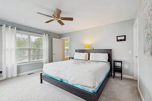 Un dormitorio con cama, ventilador de techo, mesita de noche, ventana con cortinas y puerta.