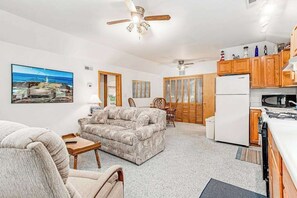 A living room with a sofa, a dining table with chairs, a kitchen area with cabinets and appliances, and a ceiling fan.