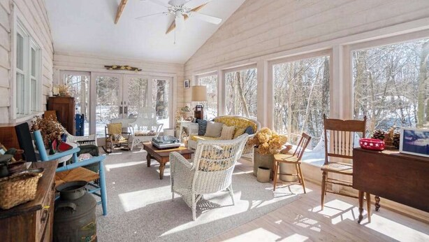 Una habitación iluminada por el sol, con ventilador de techo, ventanas amplias, muebles de mimbre y vista a los árboles.