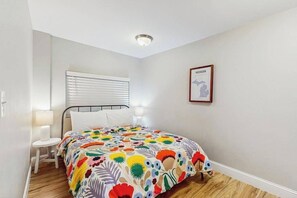 Une chambre à coucher avec un lit, une table de chevet, une lampe, une fenêtre avec des stores et un tableau encadré au mur.