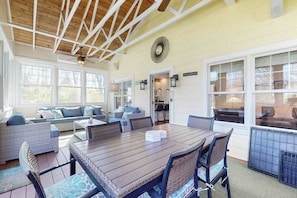 A sunlit room with a wooden ceiling, large windows, a dining table, and comfortable seating.