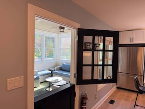 A kitchen with a stainless steel refrigerator, a dining area with a table and chairs, and a view through a window to an outdoor seating area.