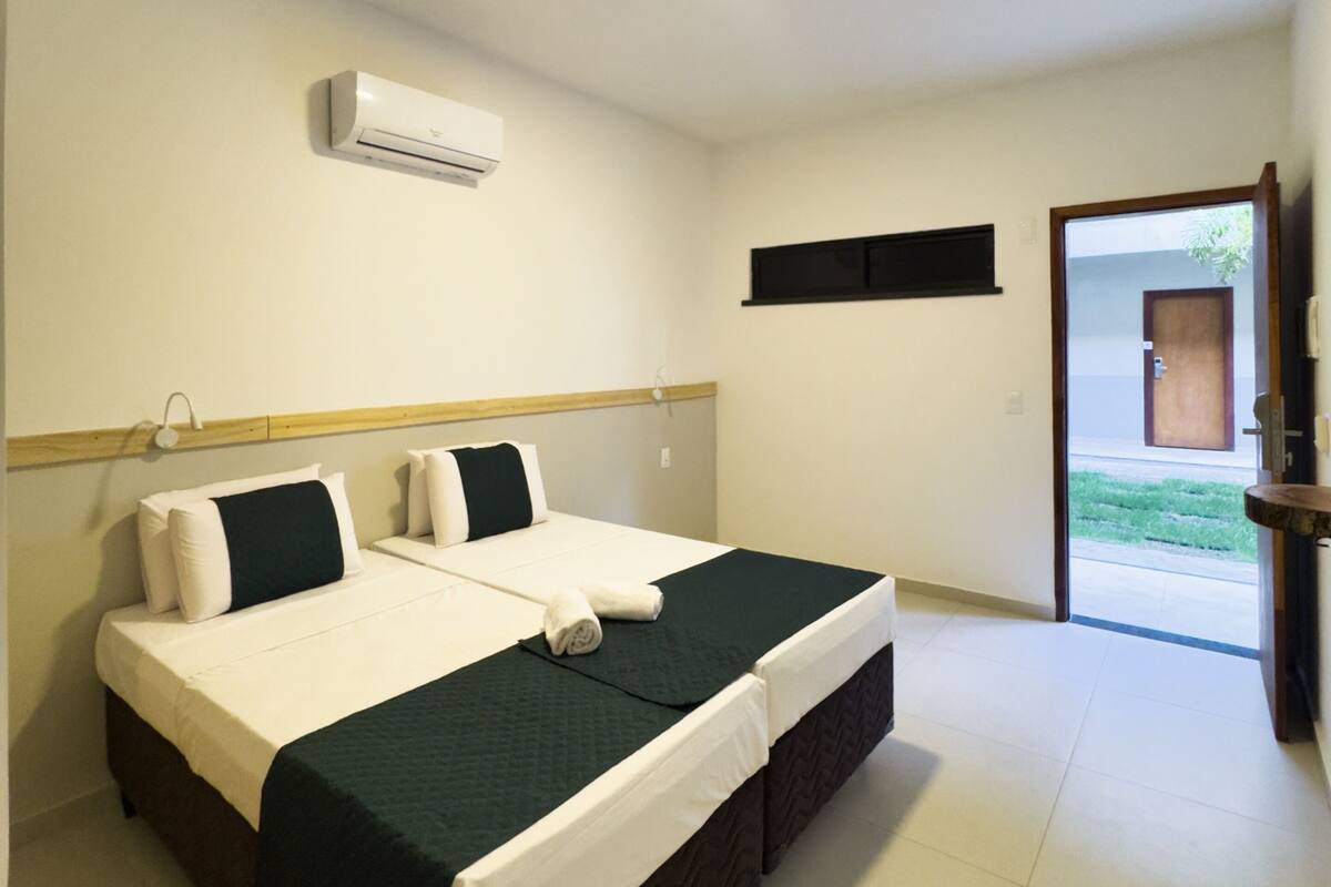 Una cama doble con colcha verde oscuro, almohadas blancas y edredón blanco. Se ve una ventana pequeña con marco negro, un equipo de aire acondicionado blanco montado en la pared y una puerta que da al exterior.