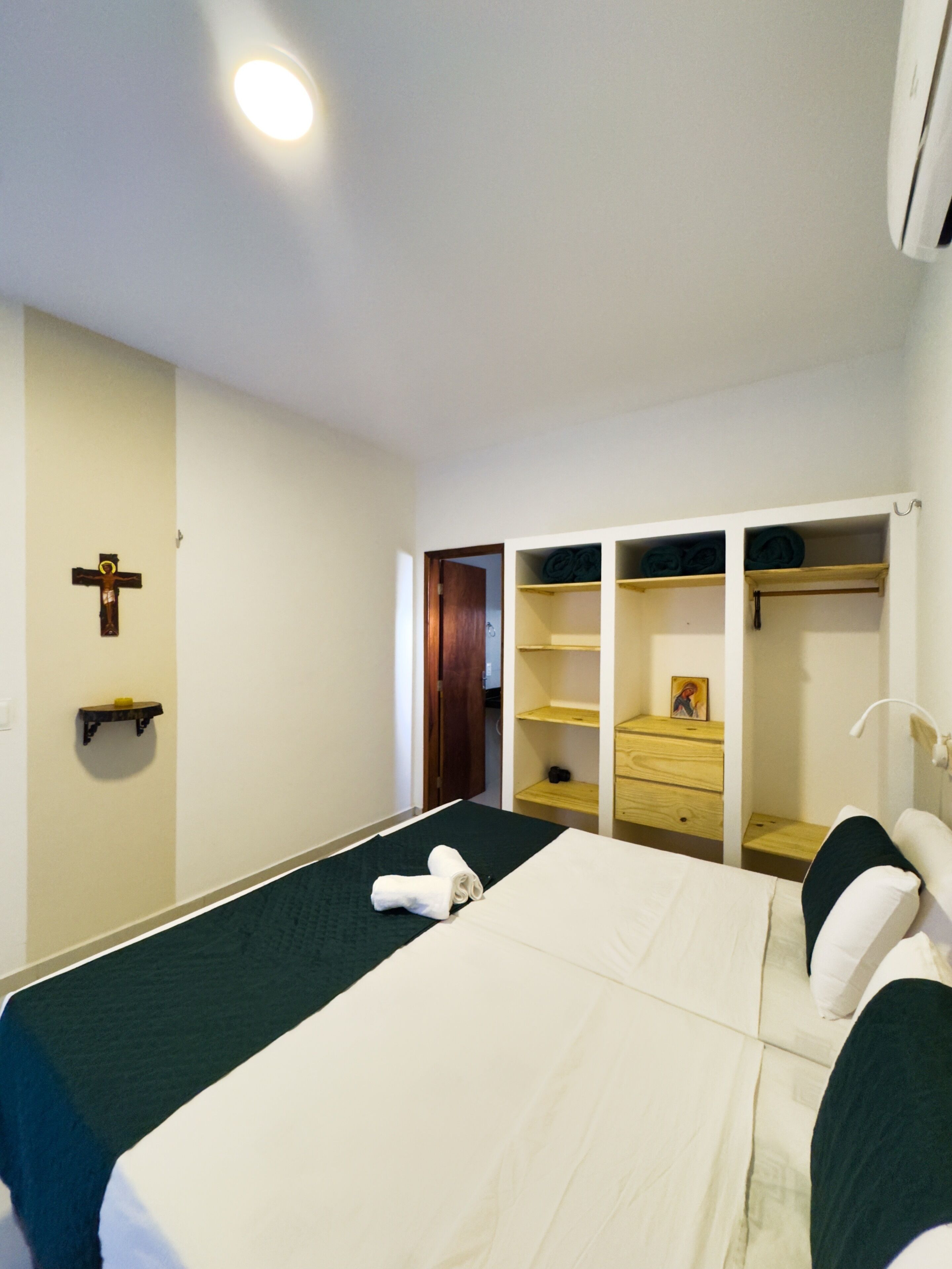 Ein Hotelzimmer mit Bett, einem Holzkreuz an der Wand, einem Kleiderschrank mit Regalen und einem kleinen Ablagetisch mit Kerze.