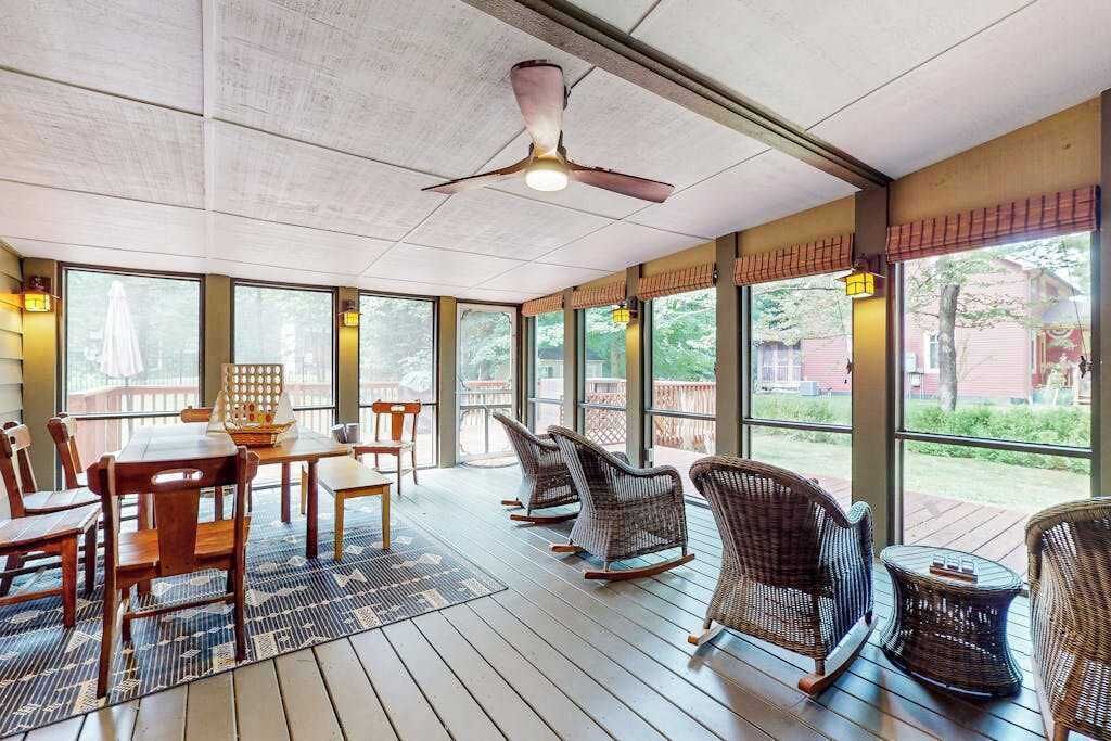 Un patio cubierto con mesa y sillas, mecedoras de mimbre y ventanas amplias con vistas al exterior.