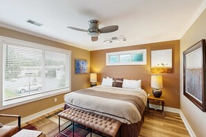 A bedroom with a large bed, a ceiling fan, two bedside lamps, a bench, and artwork on the walls.