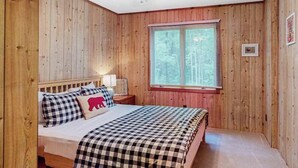 Un dormitorio con paredes de madera, una cama con ropa de cuadros, una mesita de noche con una lámpara y una ventana con persianas.