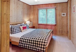 A bedroom with wooden panel walls, a bed with checkered bedding, a nightstand with a lamp, and a window with blinds.