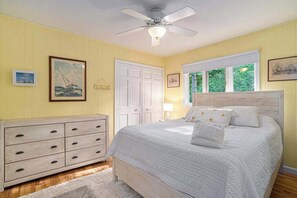 Une chambre à coucher comprenant un lit, une commode, un ventilateur de plafond et une fenêtre donnant sur un paysage verdoyant.