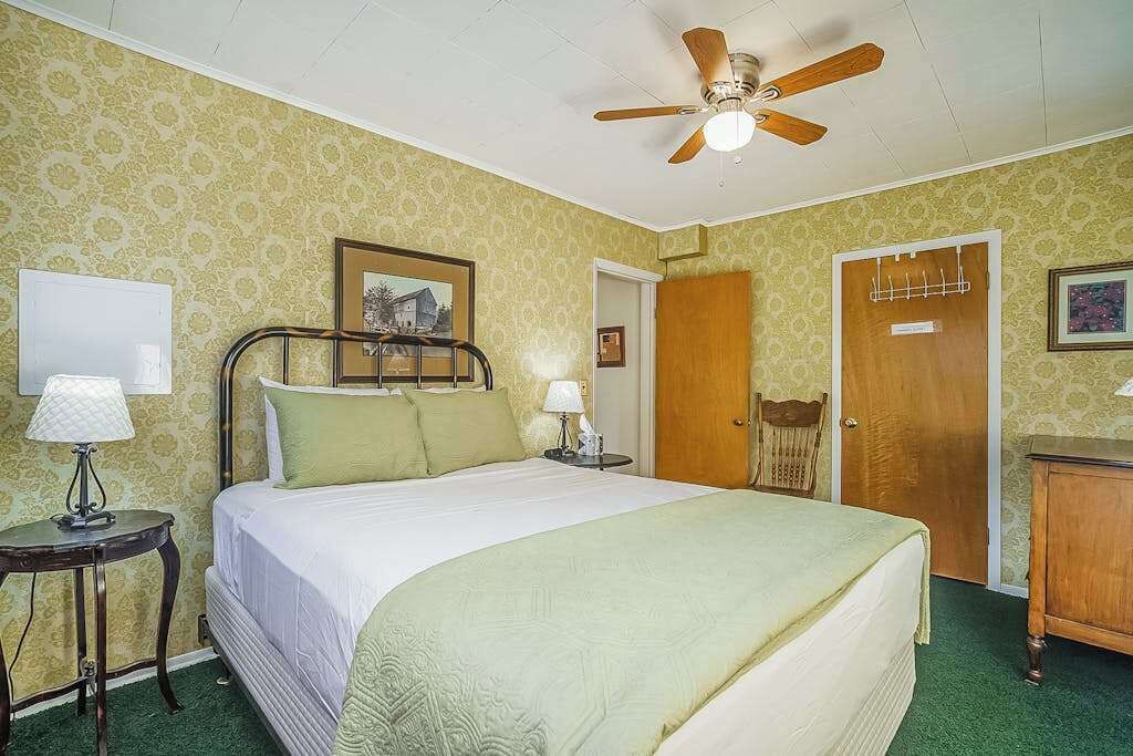 Ein Schlafzimmer mit einem Bett, einem Deckenventilator, einer Holztür, einem kleinen Tisch mit Lampe und einem Bild an der Wand.