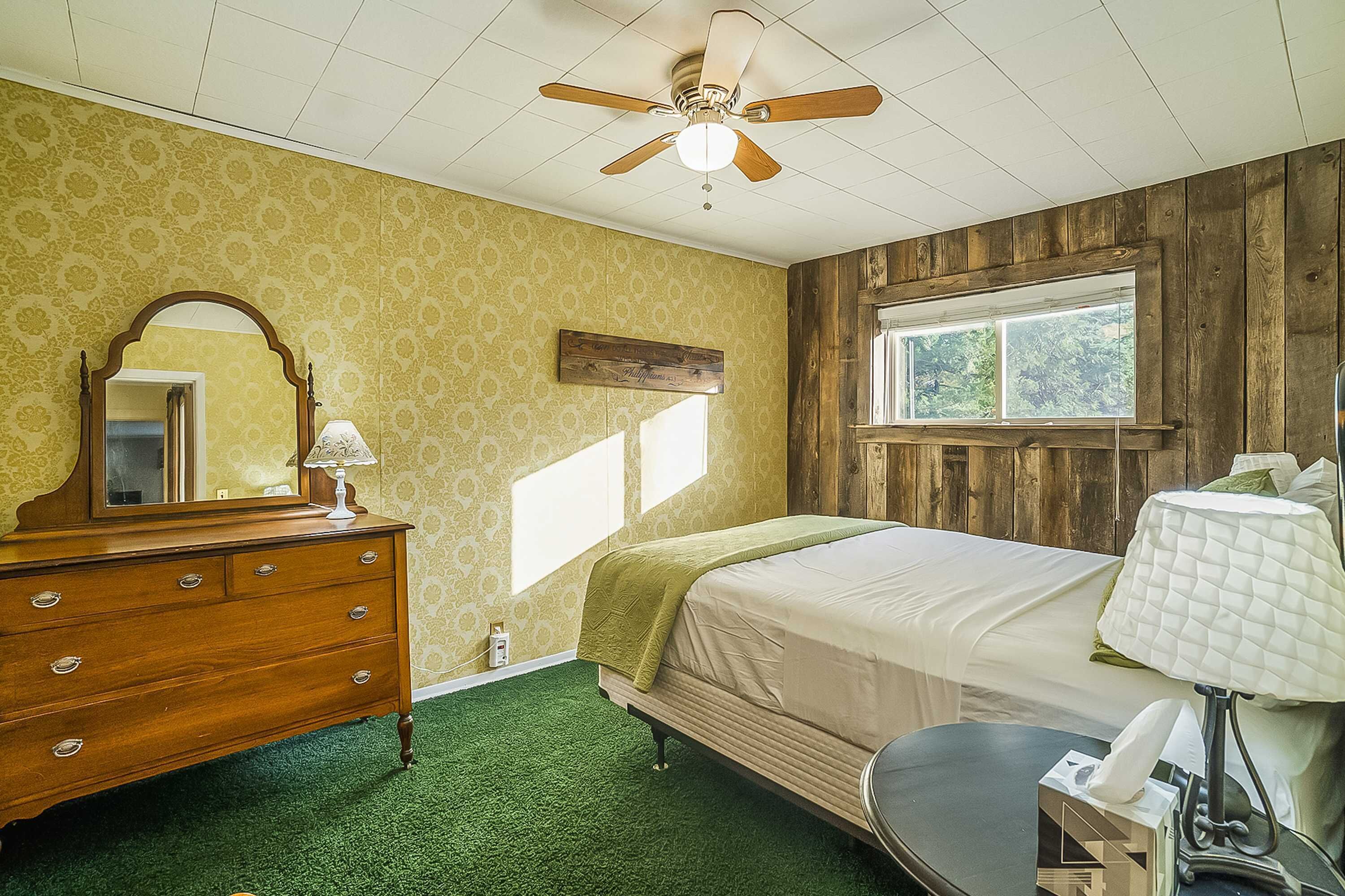 Une chambre avec une commode en bois, un lit recouvert d’une couette blanche, un ventilateur de plafond et une fenêtre donnant sur des arbres.