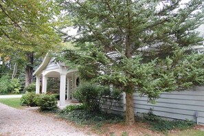 A house with a covered porch, a large tree in front, and a well-maintained garden. - Charming home near Glen Arbor with a porch, gas grill, lake views, & AC (Maple City)