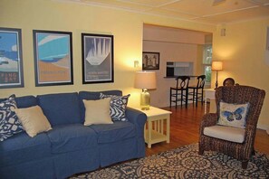 A living room with a blue sofa, a patterned armchair, a small table with a lamp, and framed pictures on the wall. - Charming home near Glen Arbor with a porch, gas grill, lake views, & AC (Maple City)