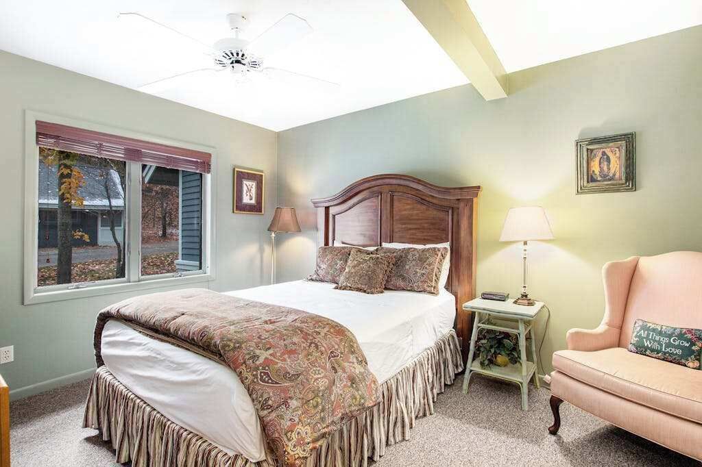 A bedroom with a bed, a nightstand, a chair, a window with blinds, and a ceiling fan.