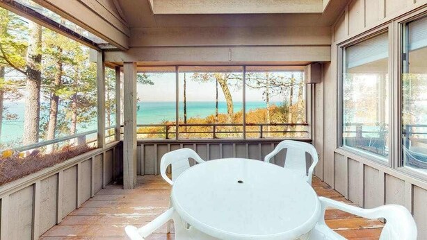 Een overdekt terras met een tafel en stoelen, grote ramen en uitzicht op de oceaan.