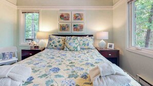 Une chambre à coucher avec une couvre-lit à motifs floraux, deux lampes de chevet, une table de chevet avec une lampe, un tableau encadré au-dessus du lit et une fenêtre donnant sur des arbres.