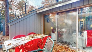 A covered outdoor dining area with a table and chairs, a sliding glass door leading to an interior space, and a wooden deck with fallen leaves. - River-view townhouse with Lake Michigan access, deck, fireplace, and cable (Glen Arbor)