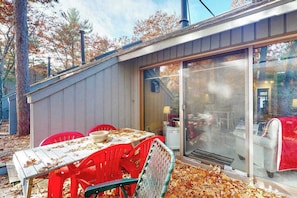 A covered outdoor dining area with a table and chairs, a sliding glass door leading to an interior space, and a wooden deck with fallen leaves.
