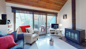A cozy living room with a sofa, TV, fireplace, and large windows. - River-view townhouse with Lake Michigan access, deck, fireplace, and cable (Glen Arbor)