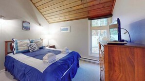 A bedroom with a wooden ceiling, a bed with blue bedding, a bedside table with a lamp, and a dresser. - River-view townhouse with Lake Michigan access, deck, fireplace, and cable (Glen Arbor)