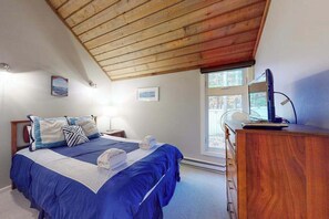 A bedroom with a wooden ceiling, a bed with blue bedding, a bedside table with a lamp, and a dresser.