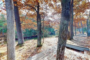 A wooden deck surrounded by trees with autumn leaves, a small bridge, and a pathway leading to a gazebo.