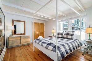 Ein Himmelbett mit kariertem Überwurf, eine hölzerne Kommode, ein Spiegel und ein Fenster mit Blick auf Gebäude.