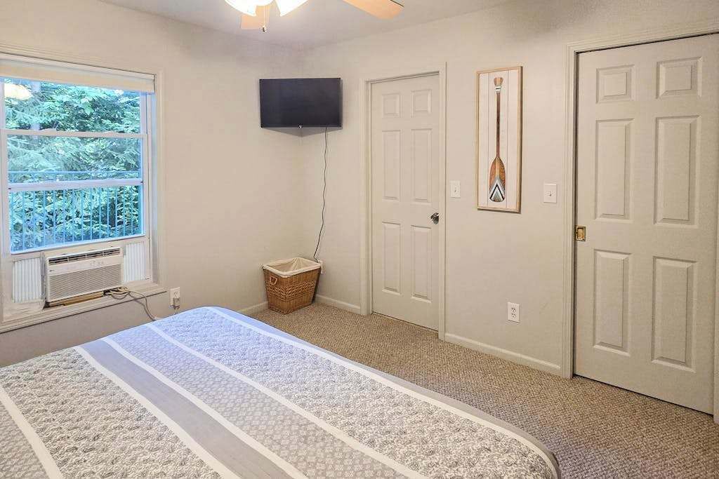 A bedroom with a bed, a television, a door, and a window with a view of trees.