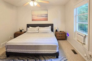 A bedroom with a bed, nightstands, a ceiling fan, a window with a view, and a wall-mounted air conditioner.